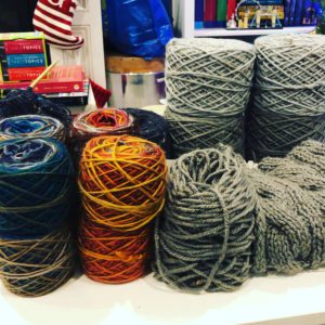 14 yarn cakes of various colors stacked around a partially used cake and a scarf in progress