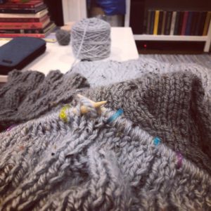 close up of a gray scarf being knit in progress with stitch markers and a yarn cake in the background