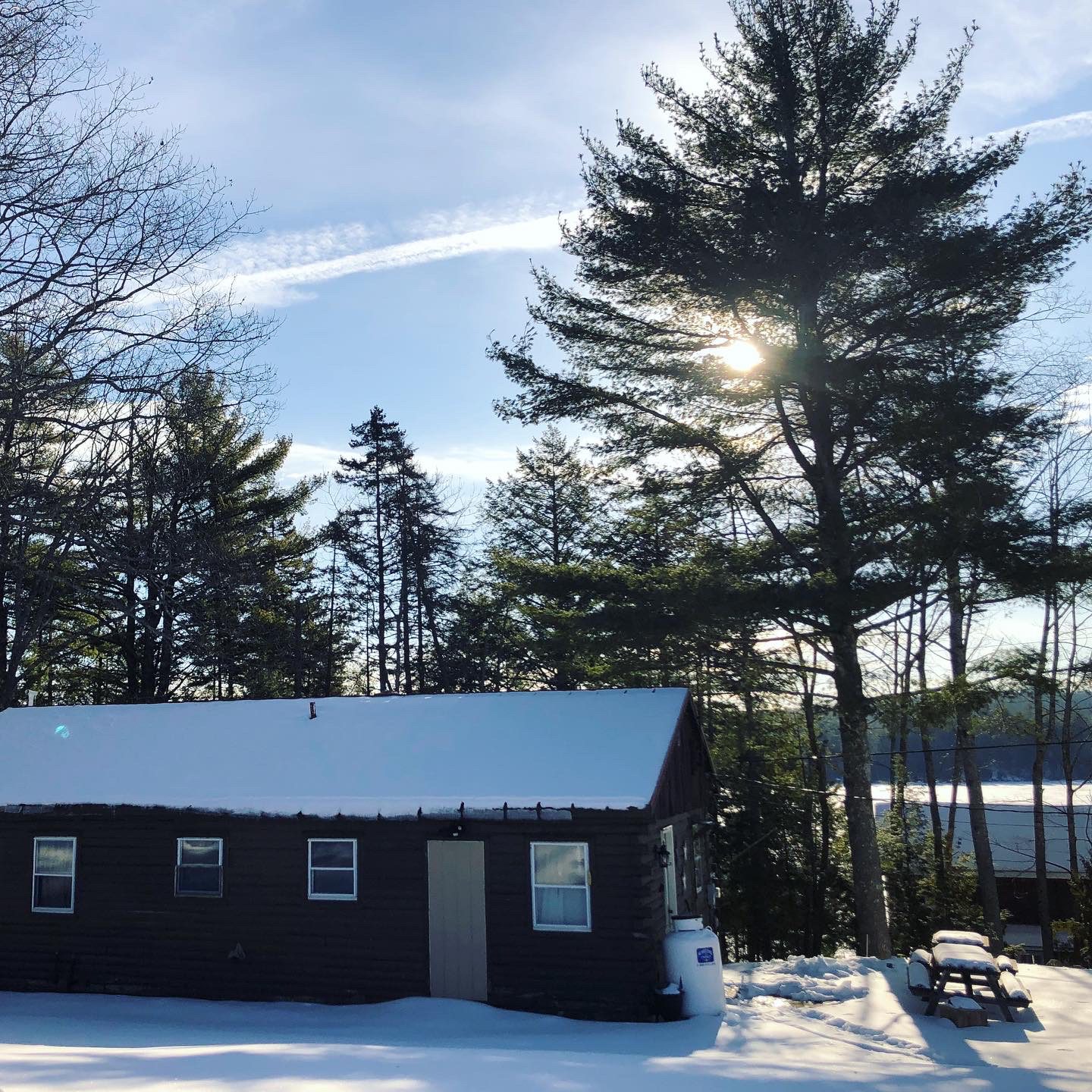 photo of a cabin covered in snow with a picnic table to the right of the cabin and a frozen lake in the background